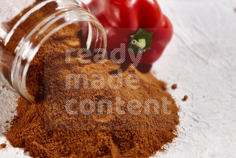 A glass jar full of ground paprika powder flipped with some spilling powder on white background