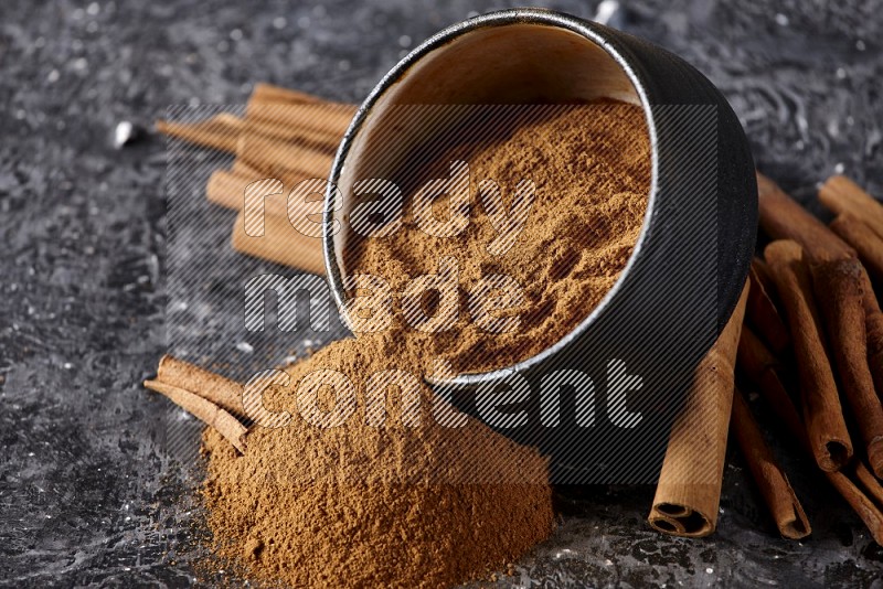 Black pottery bowl over filled with cinnamon powder and cinnamon sticks around the bowl on a textured black background