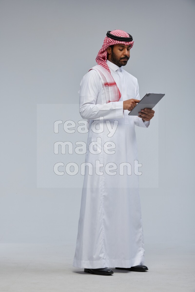 Saudi man Wearing Thob and shomag standing working on tablet on Gray background