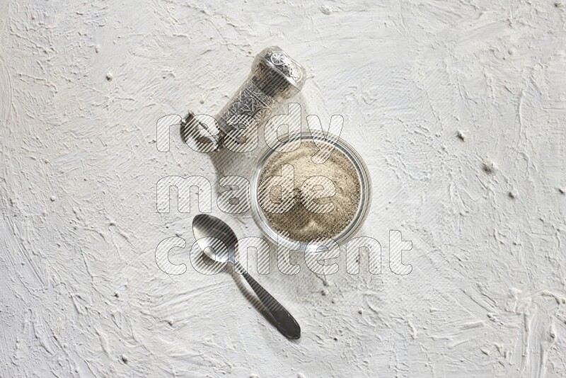 A glass bowl full of white pepper powder with white pepper beads and a metal grinder on textured white flooring