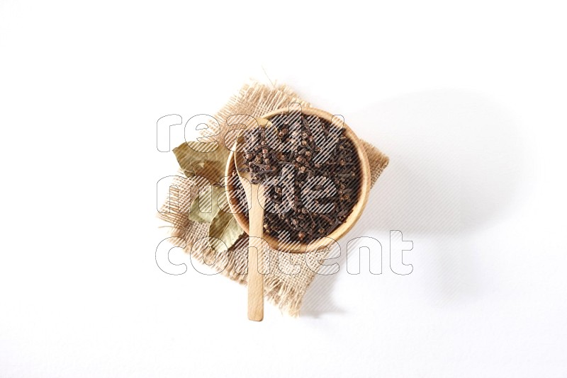 A wooden bowl and a wooden spoon full of cloves on a piece of burlap on a white flooring