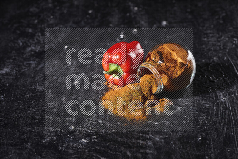 A glass jar full of ground paprika powder flipped with some spilling powder on black background