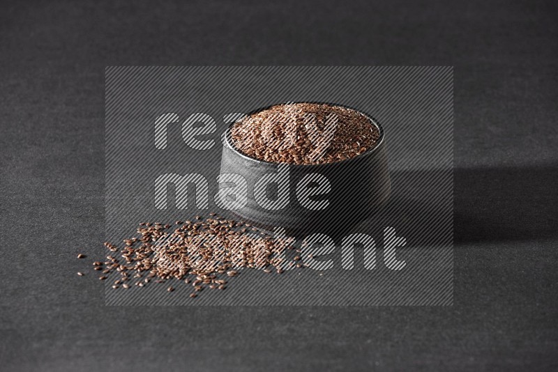 A black pottery bowl full of flax surrounded by the seeds on a black flooring in different angles