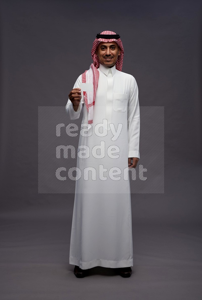 Saudi man wearing thob and shomag standing holding ATM card on gray background