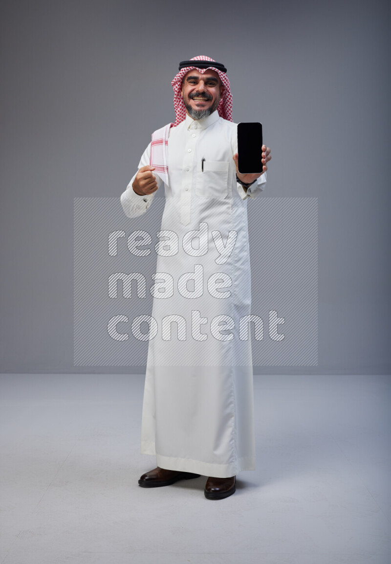 Saudi man Wearing Thob and red Shomag standing showing phone to camera on Gray background