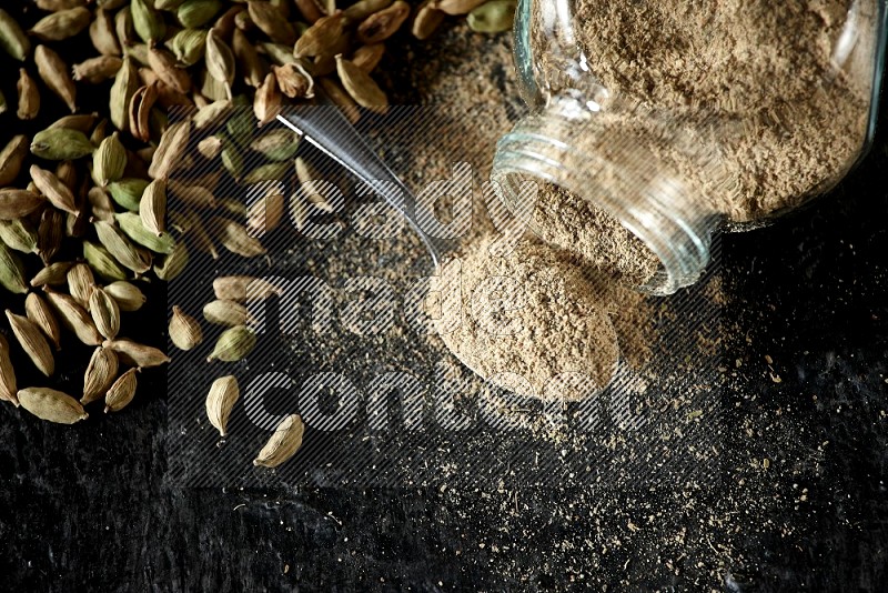 A flipped glass spice jar and a metal spoon full of cardamom powder and cardamom seeds spreaded on textured black flooring