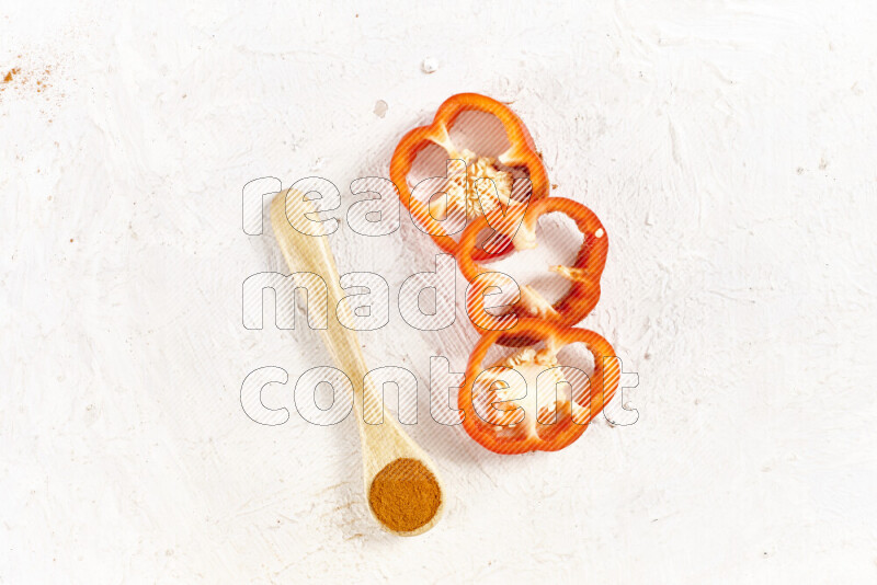 A wooden spoon full of ground paprika powder with red bell pepper slices beside it on white background