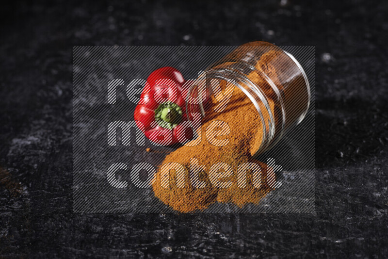 A glass jar full of ground paprika powder flipped with some spilling powder on black background