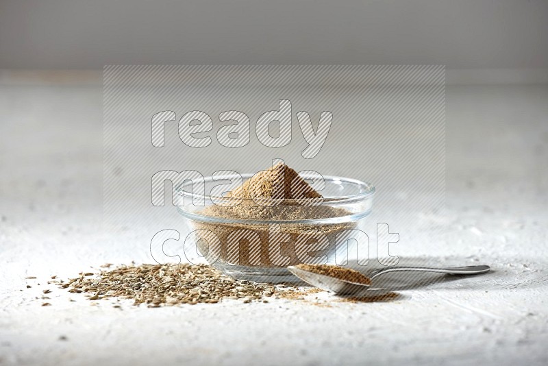A glass bowl and metal spoon full of cumin powder and cumin seeds underneath it on textured white flooring