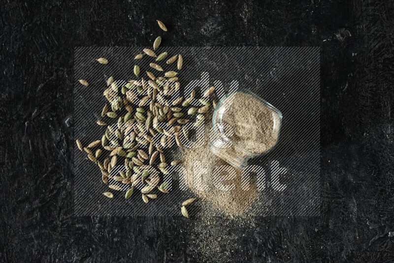 A flipped glass spice jar full of cardamom powder and powder spilled out of it with cardamom seeds spreaded on textured black flooring