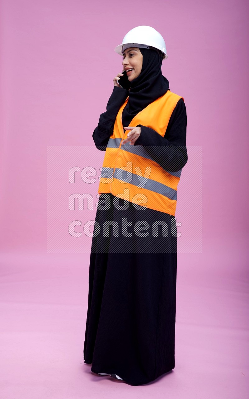 Saudi woman wearing Abaya with engineer vest and helmet standing talking on phone on pink background