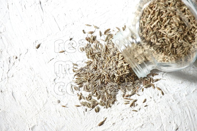 A flipped glass spice jar full of cumin seeds and the seeds spilled out on textured white flooring