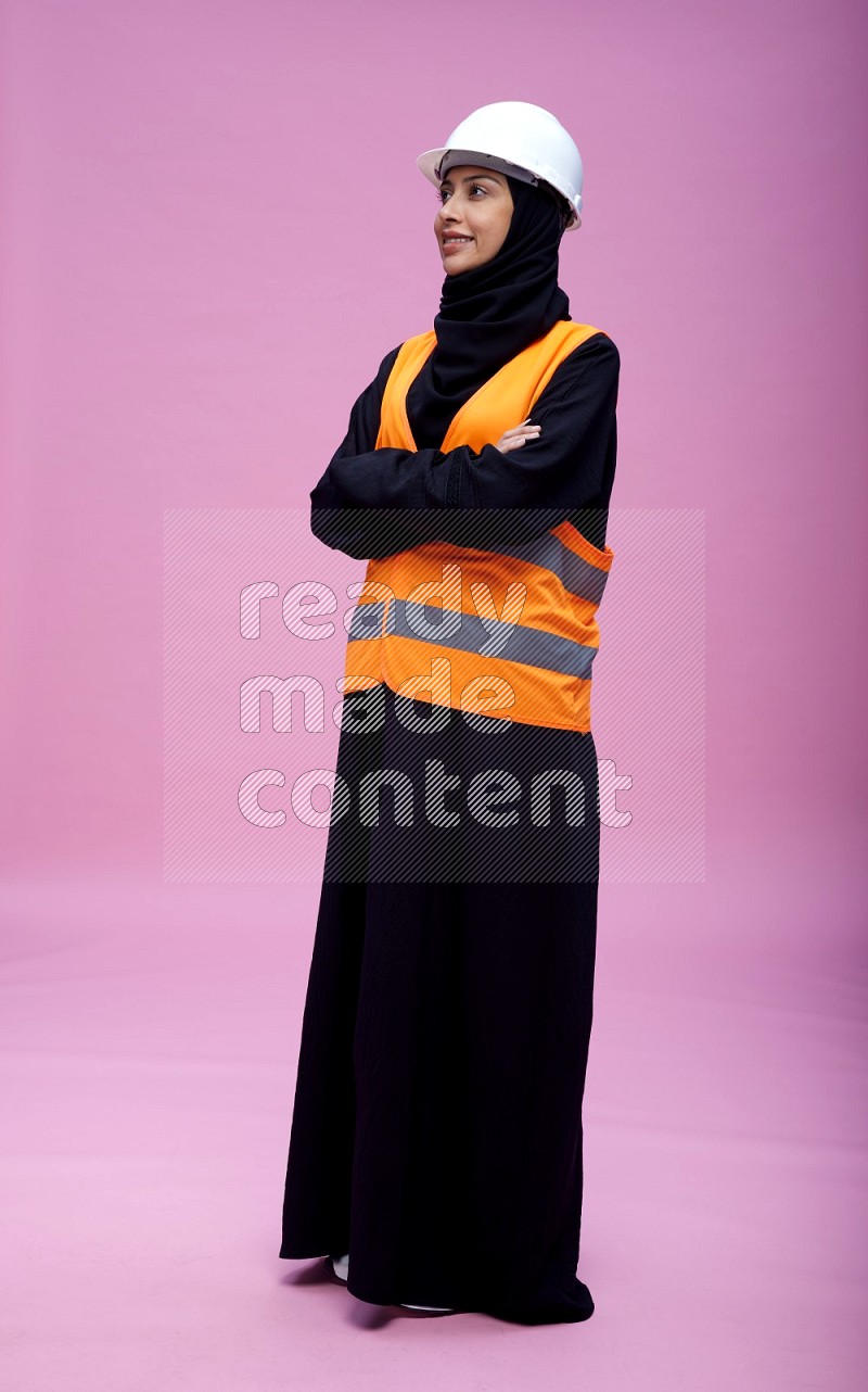 Saudi woman wearing Abaya with engineer vest and helmet standing with crossed arms on pink background