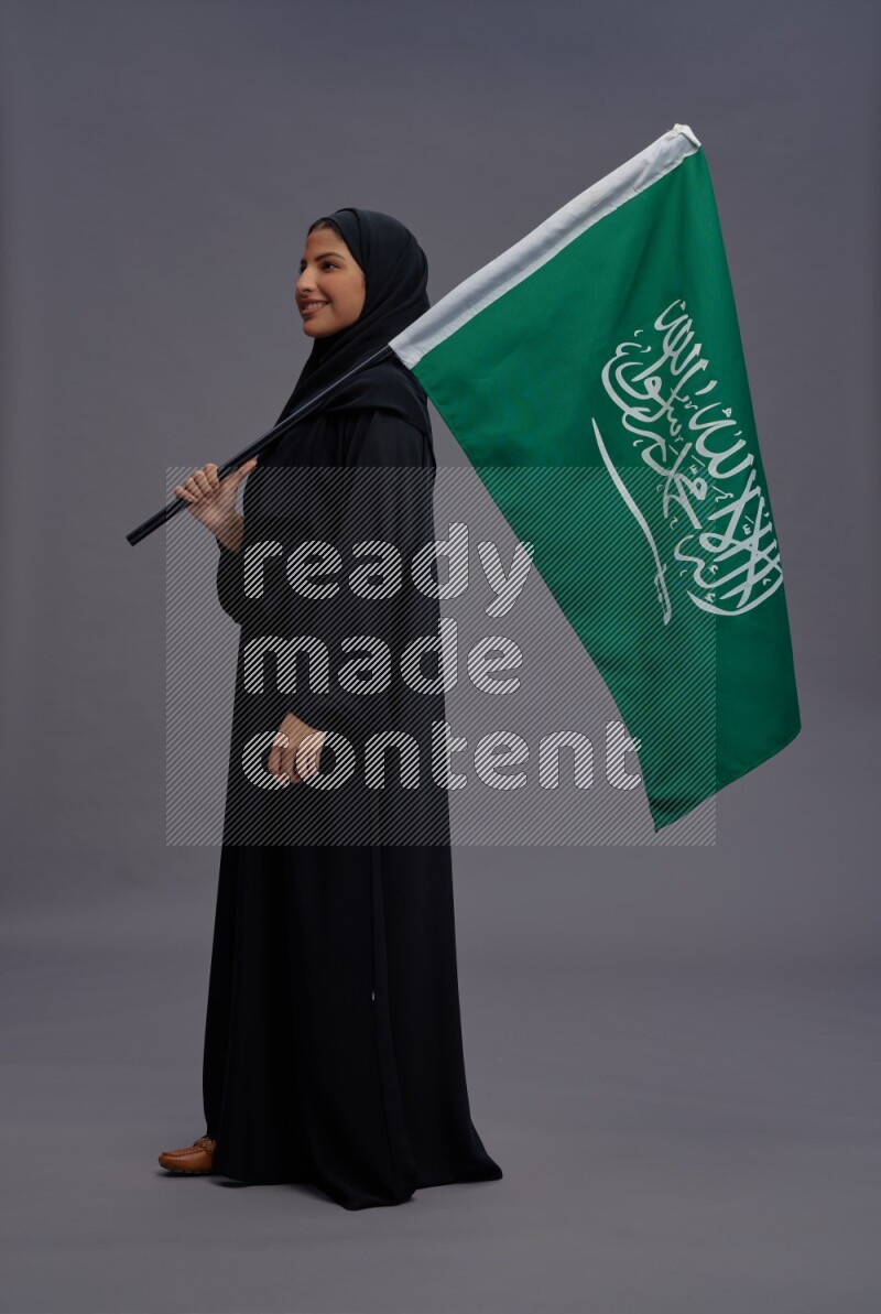 Saudi woman wearing Abaya standing holding Saudi flag on gray background