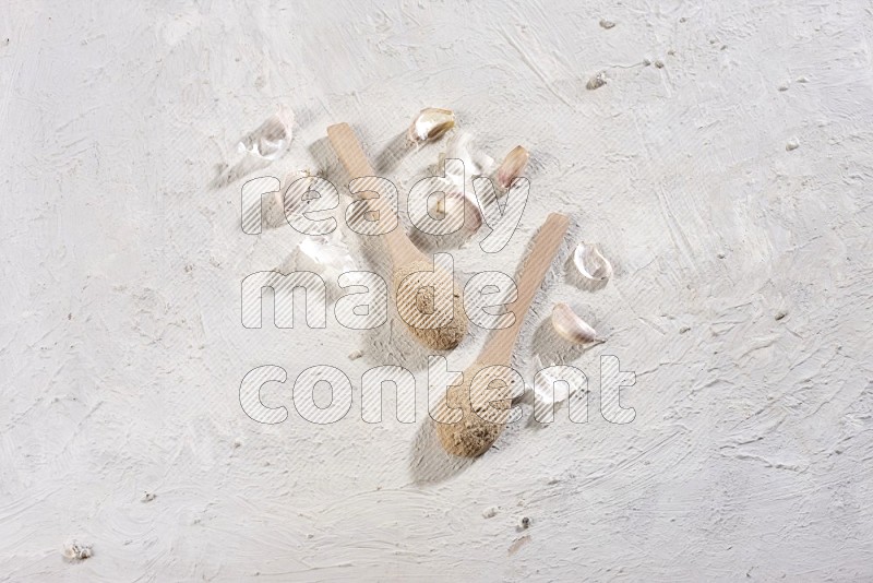 2 wooden spoons full of garlic powder with cloves and peels on a textured white flooring in different angles