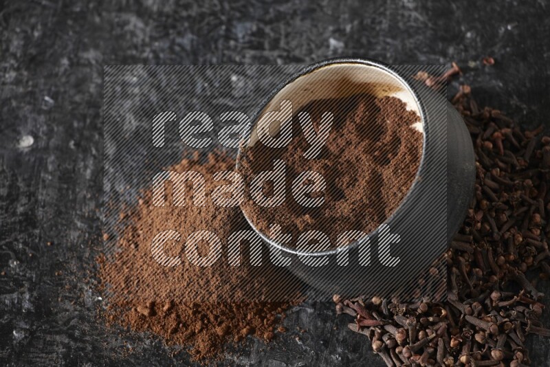 A black pottery bowl full of cloves powder spilled out of it with whole grains on a textured black flooring