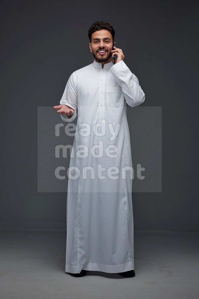 A Saudi man wearing Thobe and talking in the phone while standing and making different poses eye level on a gray background