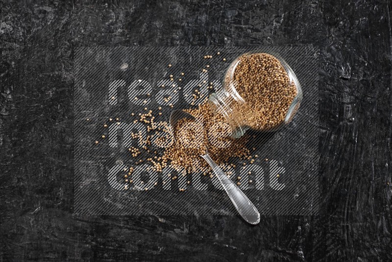 A glass spice jar and a metal spoon full of mustard seeds and jar is flipped with fallen seeds on a textured black flooring