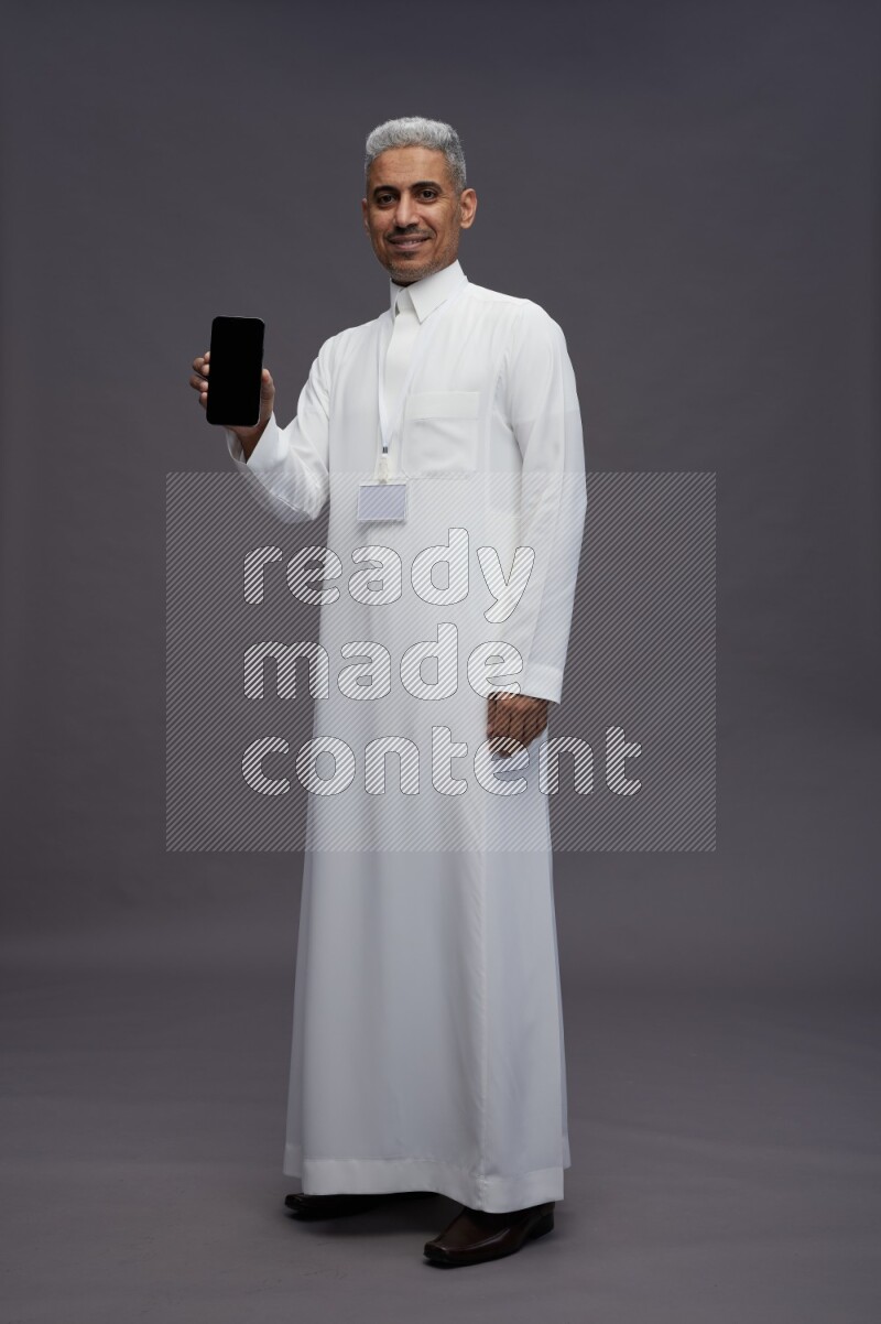 Saudi man wearing thob with neck strap employee badge standing showing phone to camera on gray background