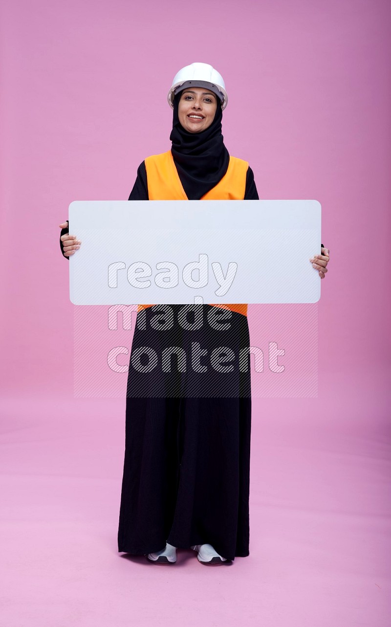 Saudi woman wearing Abaya with engineer vest and helmet standing holding board on pink background