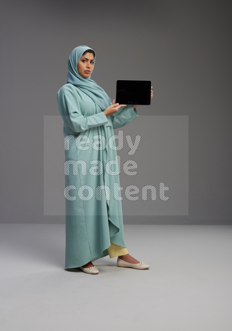 Saudi Woman wearing Abaya standing showing tablet to camera on Gray background