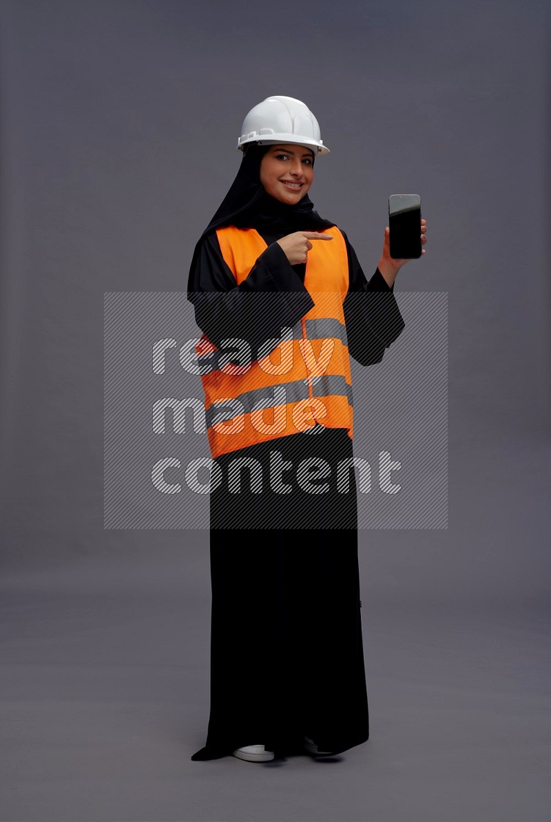 Saudi woman wearing Abaya with engineer vest standing showing phone to camera on gray background