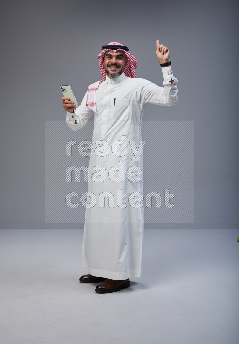 Saudi man Wearing Thob and red Shomag standing texting on phone on Gray background