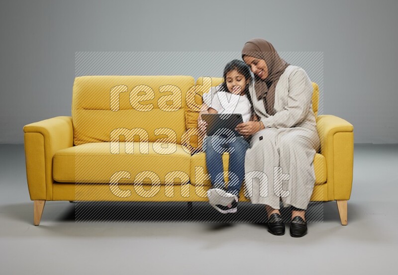 A girl with her mother on yellow sofa and watching on iPad on gray background