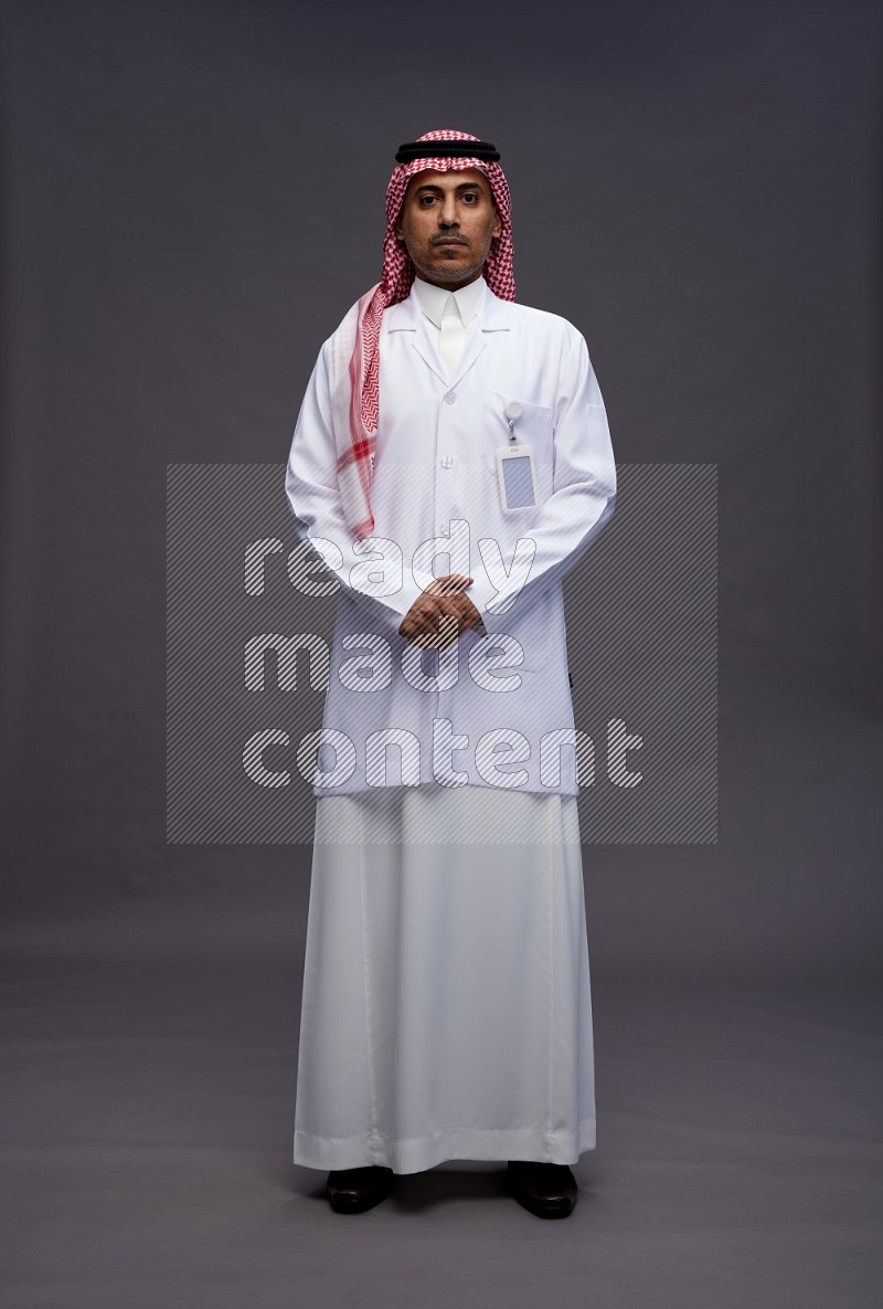 Saudi man wearing thob with lab coat and shomag with pocket employee badge standing interacting with the camera on gray background