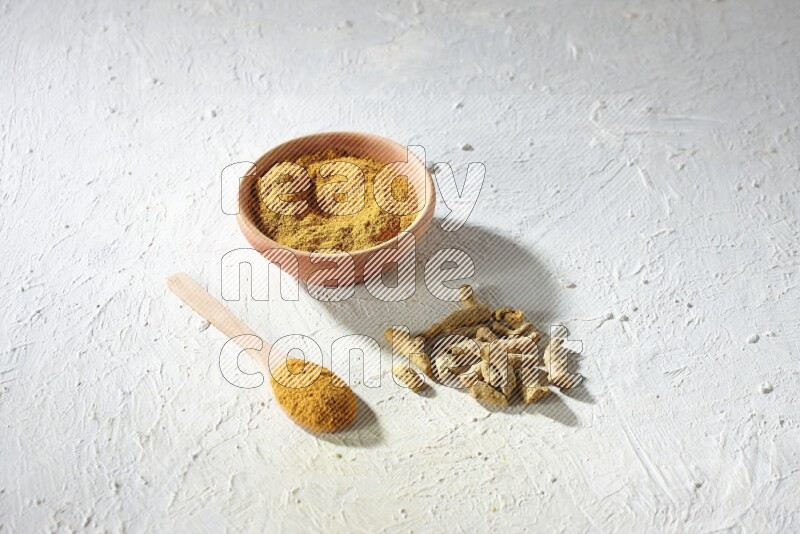 A wooden bowl and wooden spoon full of turmeric powder with dried turmeric fingers on textured white flooring