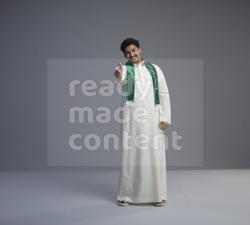 A Saudi man standing wearing thob and Saudi flag scarf on gray background