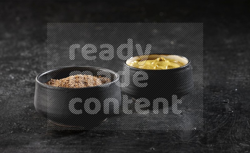 2 black pottery bowls full of mustard seeds and mustard paste on black flooring