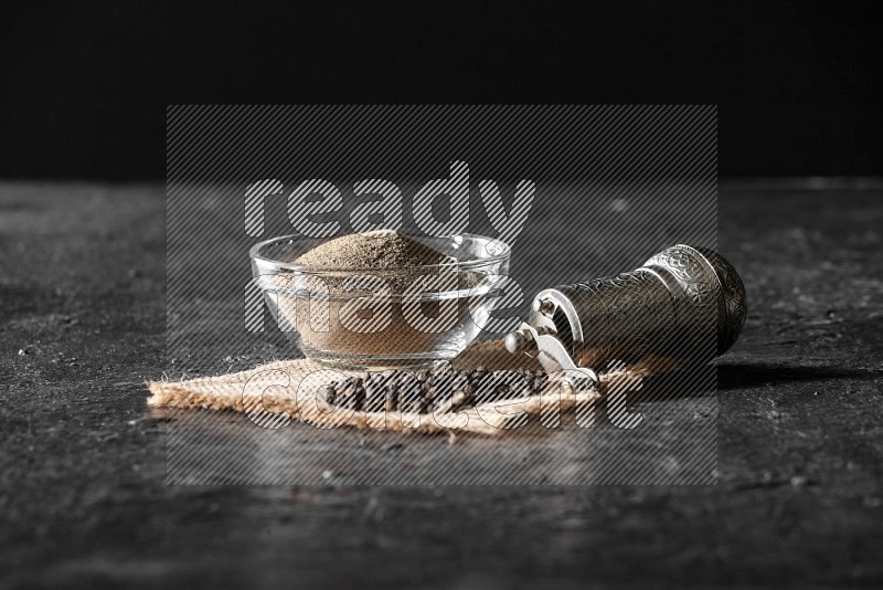 A glass bowl full of black pepper powder and black pepper beads on burlap fabric with a turkish metal pepper grinder on textured black flooring