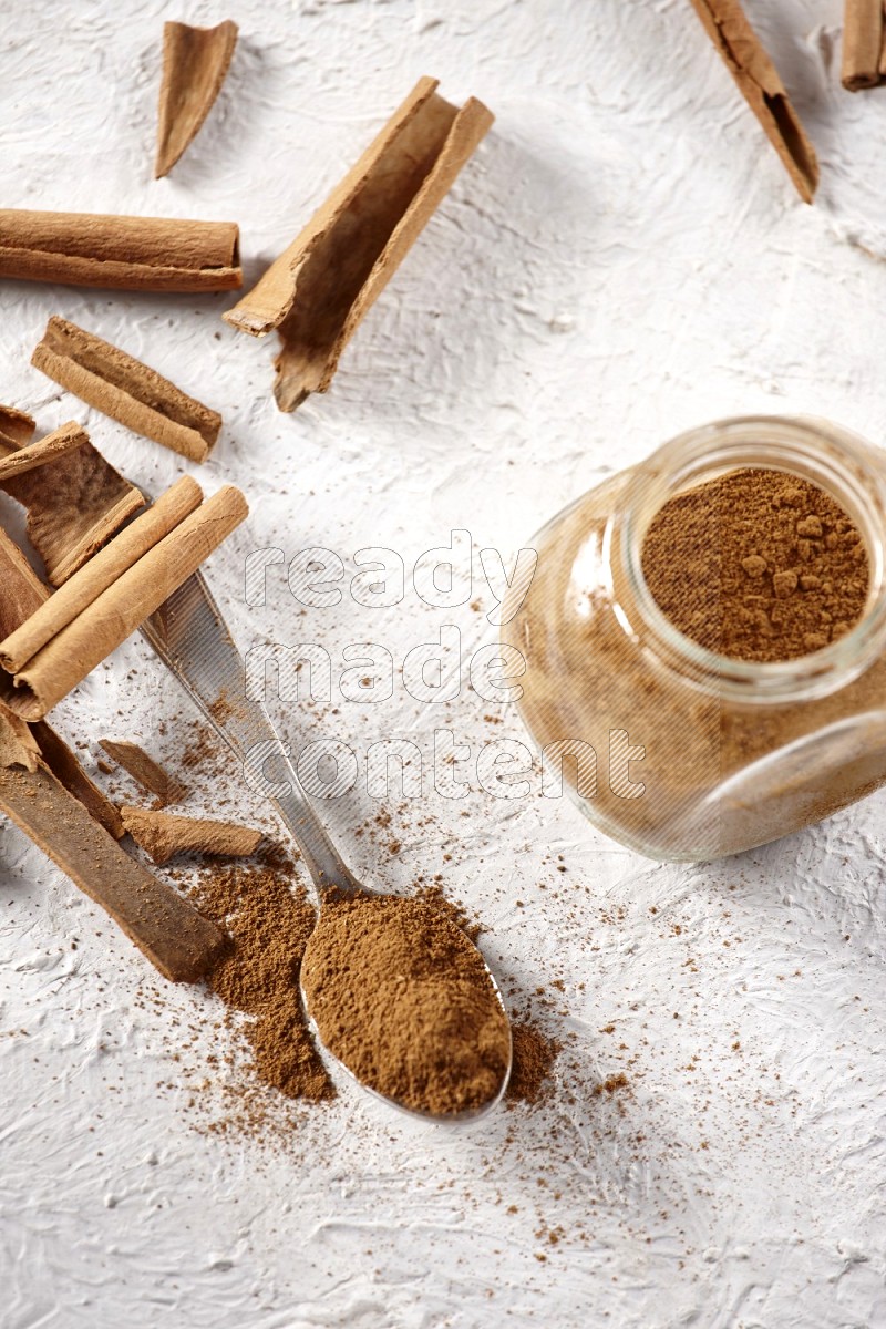 Herbal glass jar full cinnamon powder and a metal spoon surrounded by cinnamon sticks on a white background