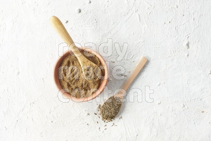 A wooden bowl and 2 wooden spoons full of cumin powder and cumin seeds on textured white flooring