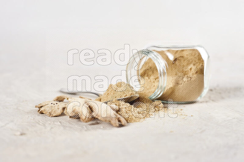 A glass jar full of ground ginger powder flipped with some spilling powder on white background