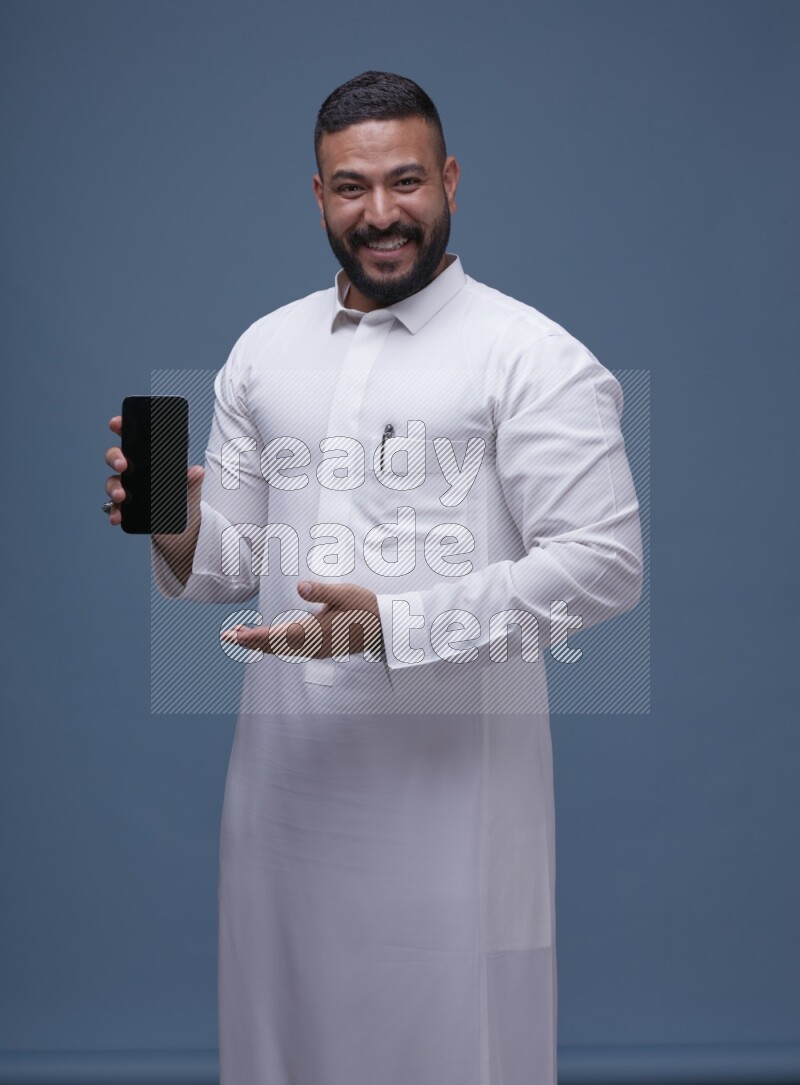 A man Showing his phone in Blue Background wearing Saudi Thob