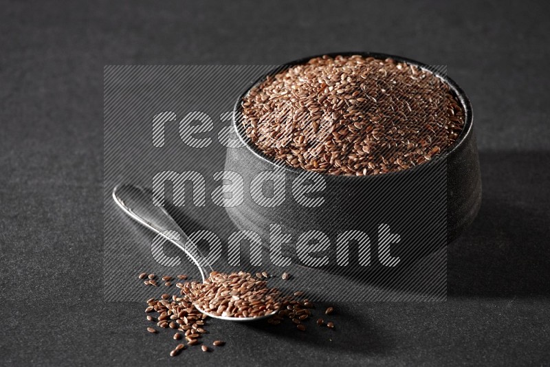 A black pottery bowl full of flax and a metal spoon full of the seeds on a black flooring in different angles