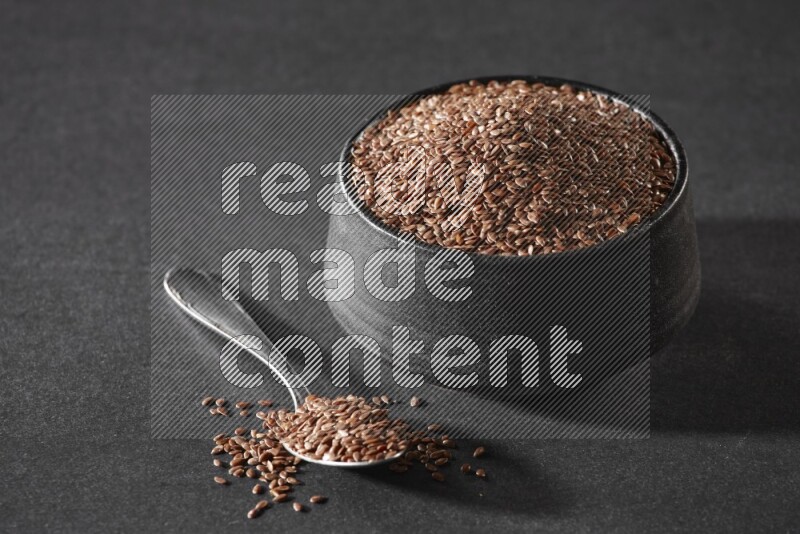 A black pottery bowl full of flaxseeds and a metal spoon full of the seeds on a black flooring