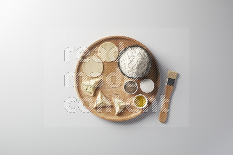 two closed sambosas and one open sambosa filled with cheese while flour, salt, black pepper and oil with oil brush aside in a wooden dish on a white background