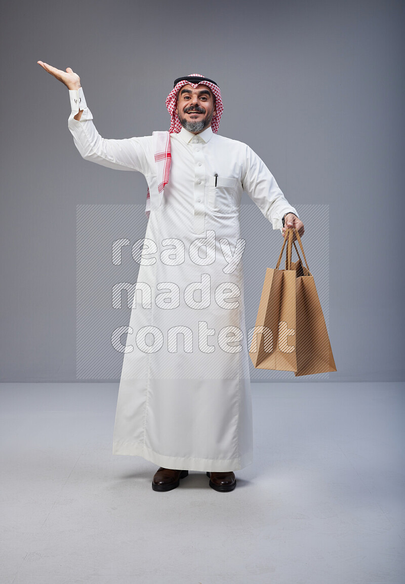 Saudi man Wearing Thob and red Shomag standing holding shopping bag on Gray background