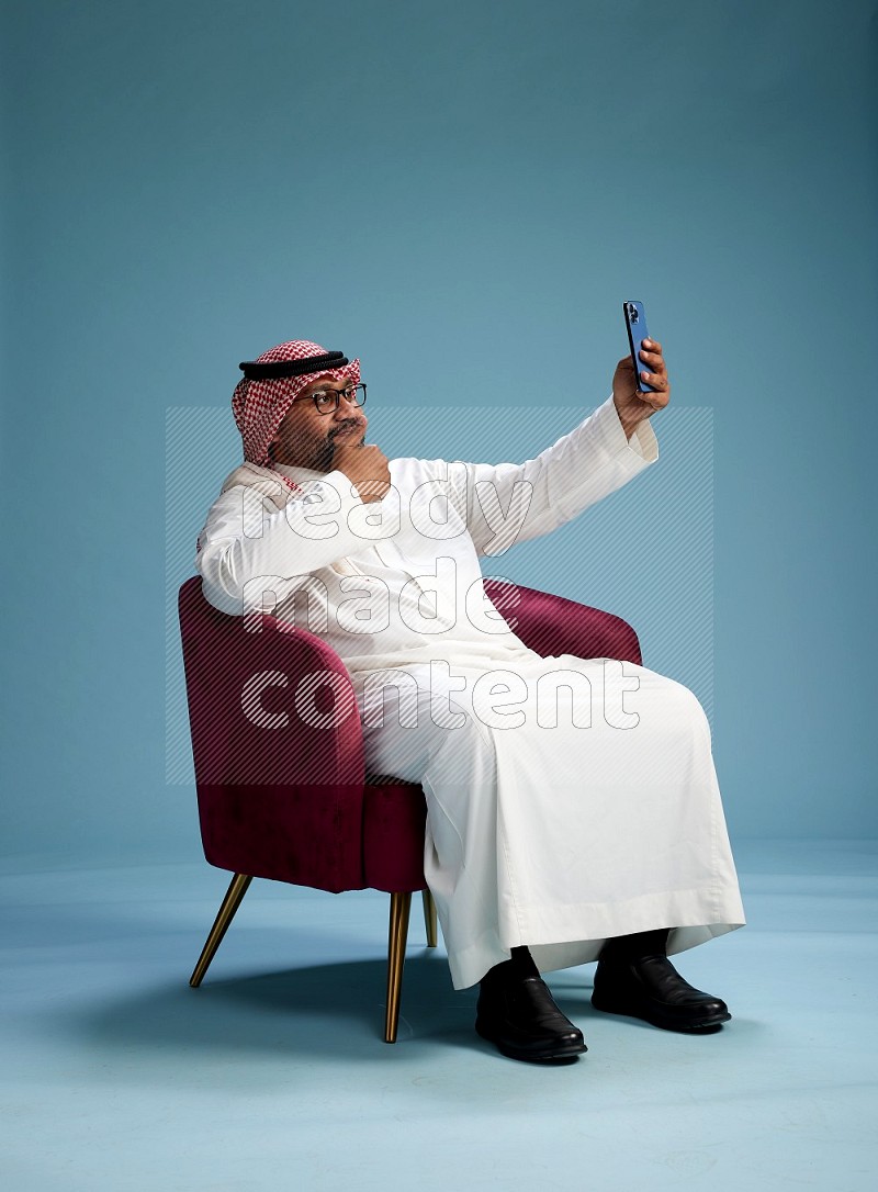 Saudi Man with shimag sitting on chair taking selfie on blue background