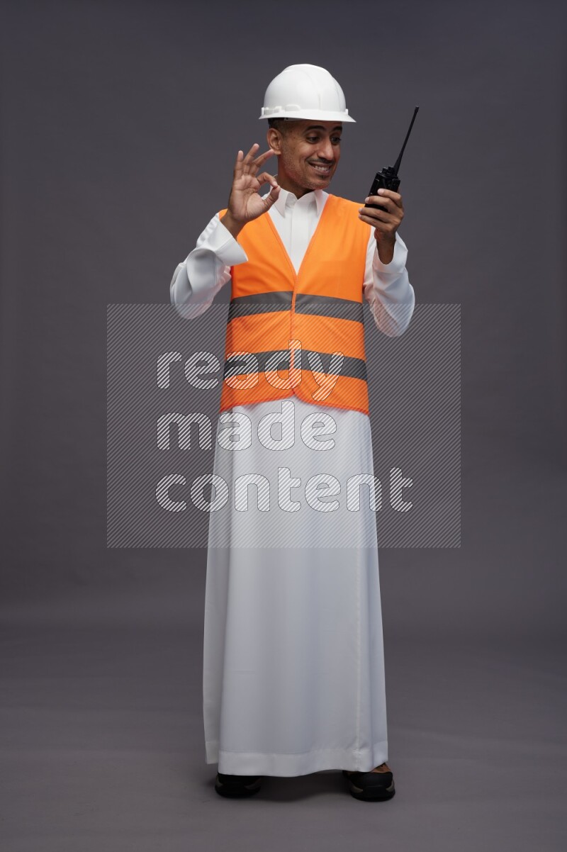 Saudi man wearing thob with engineer vest standing holding walkie-talkie on gray background