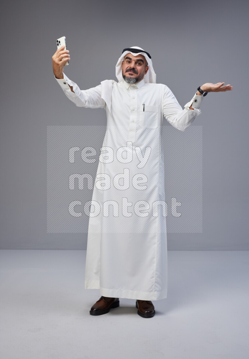 Saudi man Wearing Thob and white Shomag standing taking selfie on Gray background
