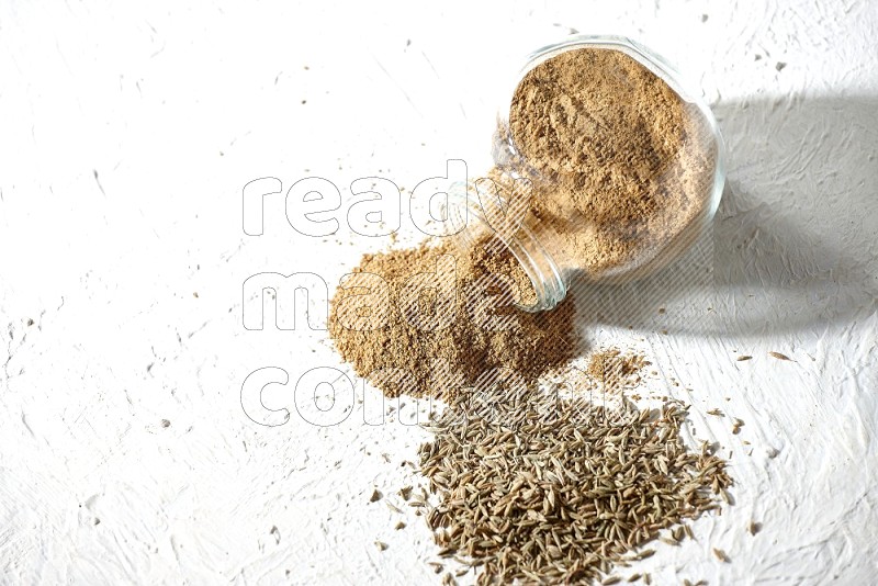 A flipped glass spice jar full of cumin powder and powder spilled out with cumin seeds on textured white flooring