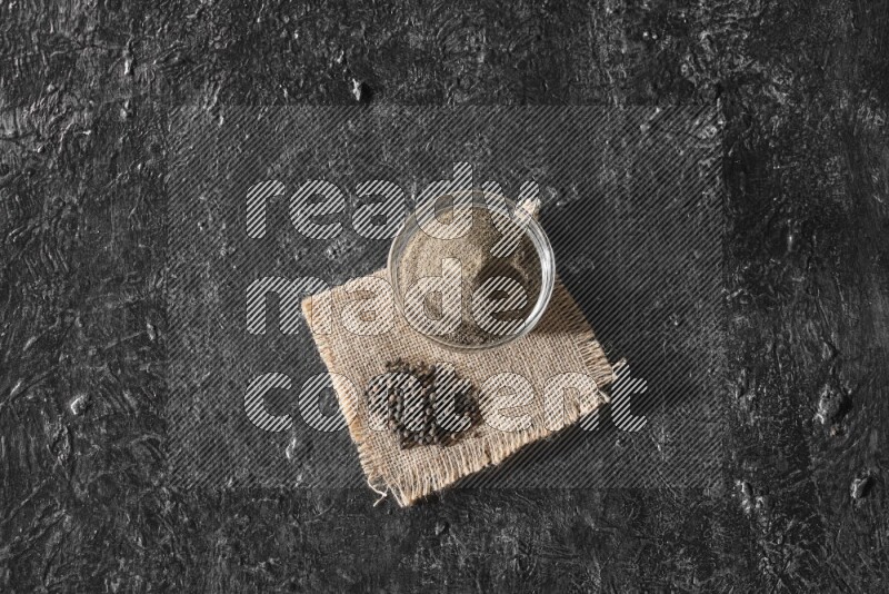 A glass bowl full of black pepper powder and black pepper beads on burlap fabric on textured black flooring