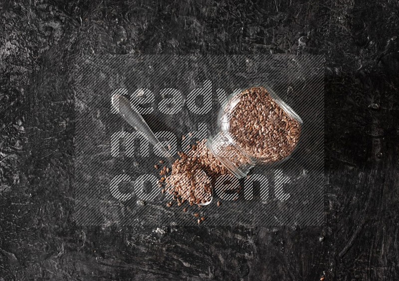 A glass spice jar full of flaxseeds flipped and seeds spread out with a metal spoon full of the seeds on a textured black flooring
