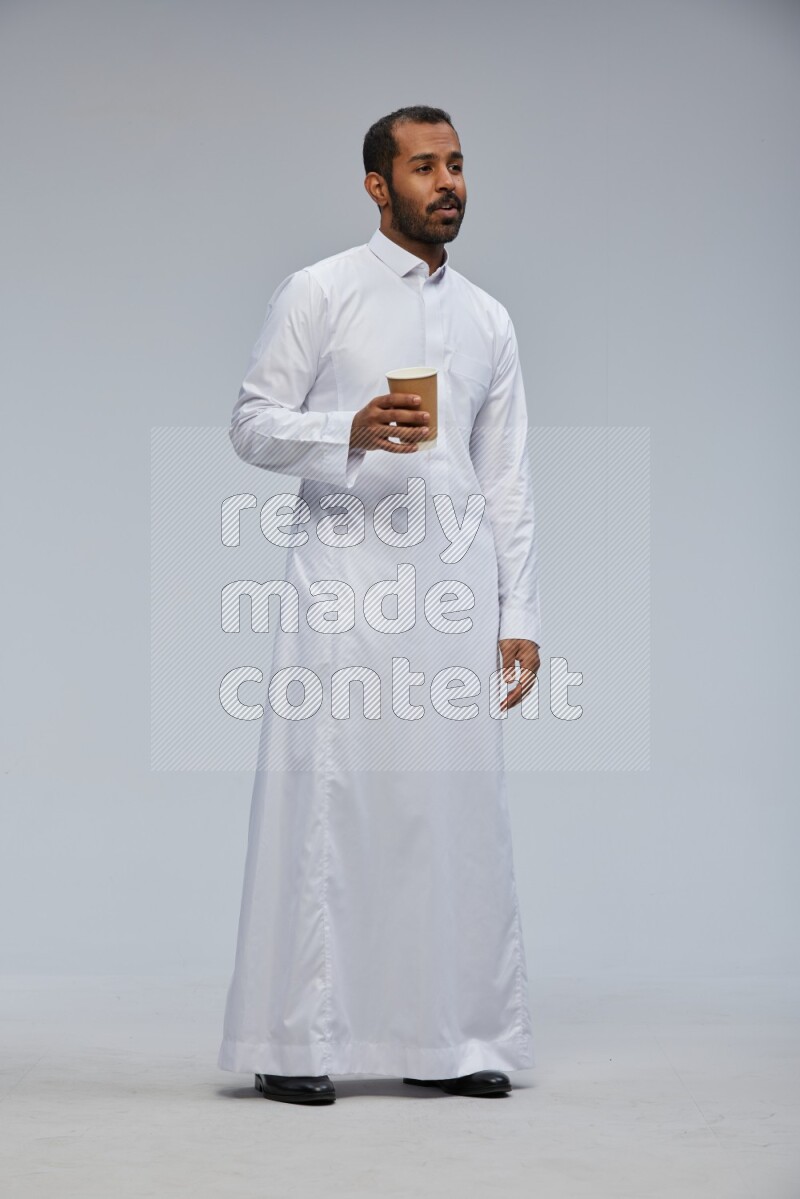Saudi man wearing thob standing holding paper cup on gray background