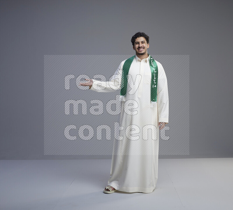 A Saudi man standing wearing thob and Saudi flag scarf on gray background