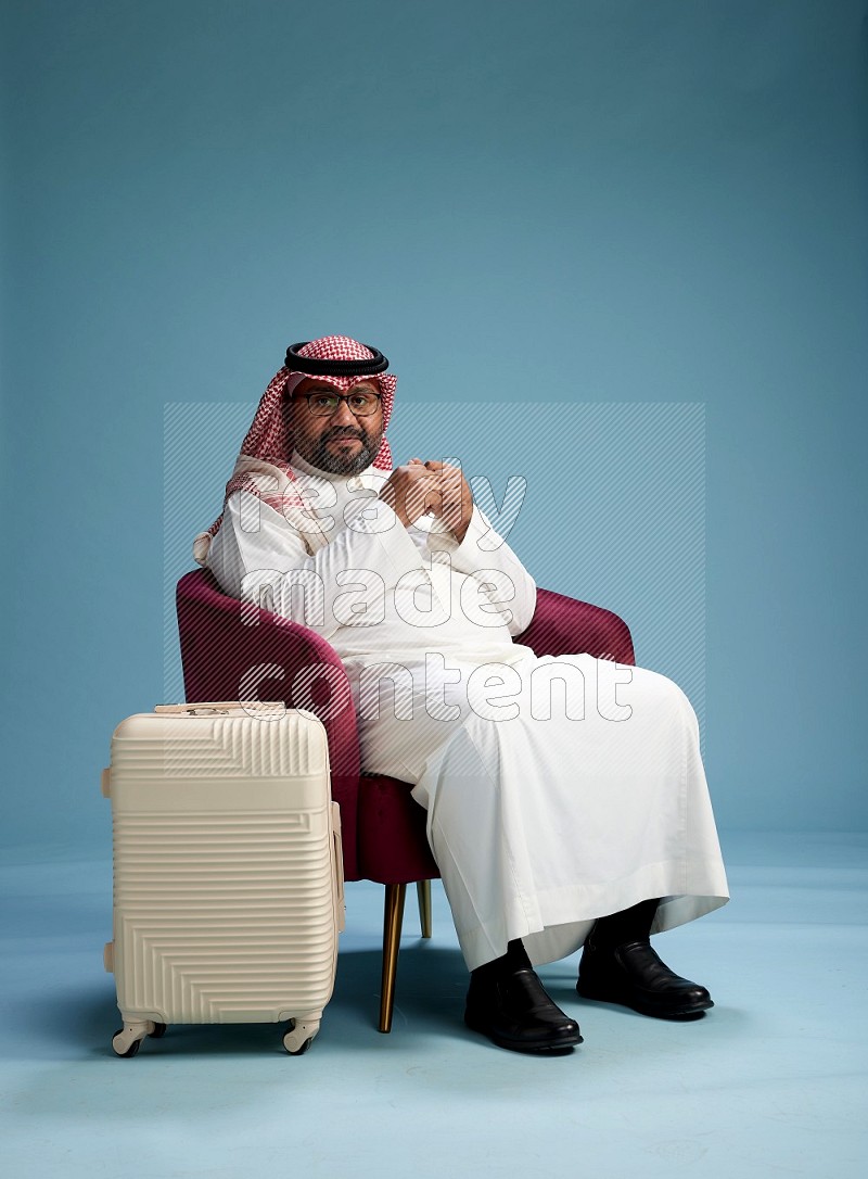 Saudi Man with shimag sitting on chair with a travel luggage on blue background